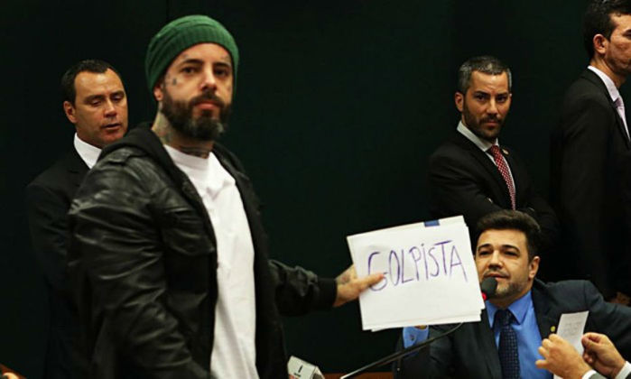 O cantor Tico Santa Cruz estende cartaz com a palavra “golpista” ao lado do deputado Marco Feliciano (PSC-SP) na Comissão da Cultura da Câmara